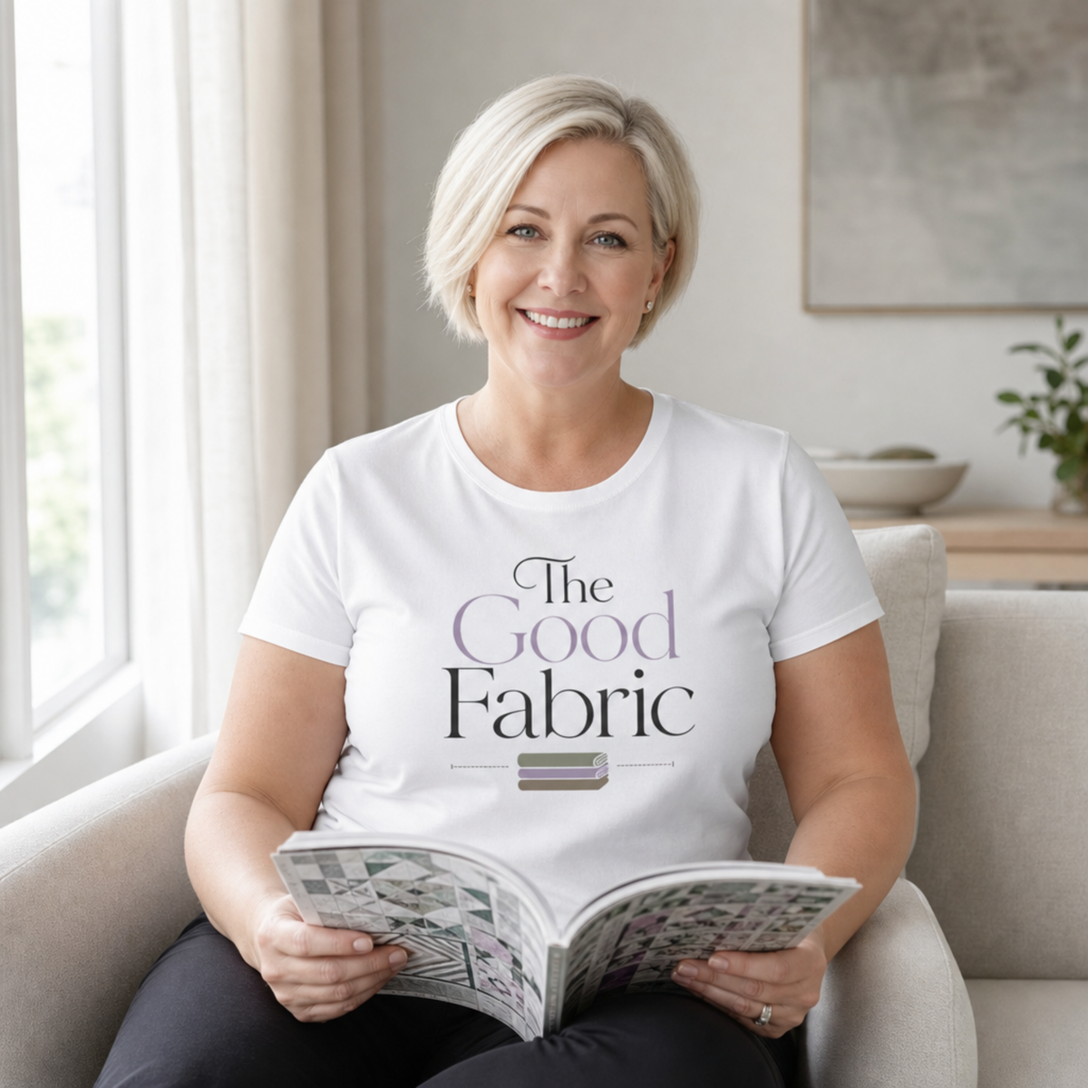 A smiling blonde woman sits in a light beige armchair holding an open book. She is wearing a white short-sleeve T-shirt with the text 'The Good Fabric' printed on the front in black and muted lavender, with a small stack of folded fabric below in muted sage, lavender and brown.