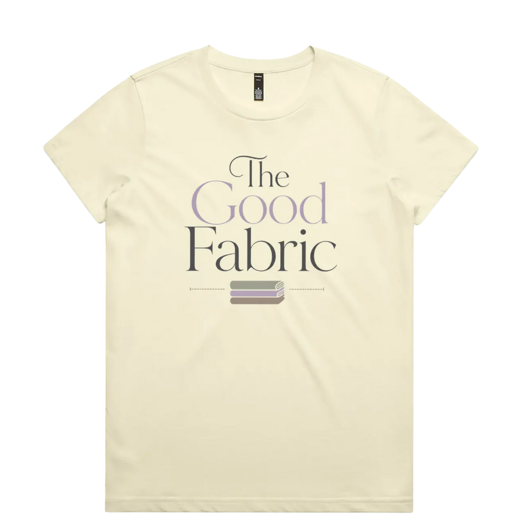 A very pale yellow short-sleeve crewneck t-shirt featuring a graphic print on the front that reads 'The Good Fabric' in dark gray and lavender lettering, with a small fabric bundle illustration below.