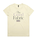 A very pale yellow short-sleeve crewneck t-shirt featuring a graphic print on the front that reads 'The Good Fabric' in dark gray and lavender lettering, with a small fabric bundle illustration below.