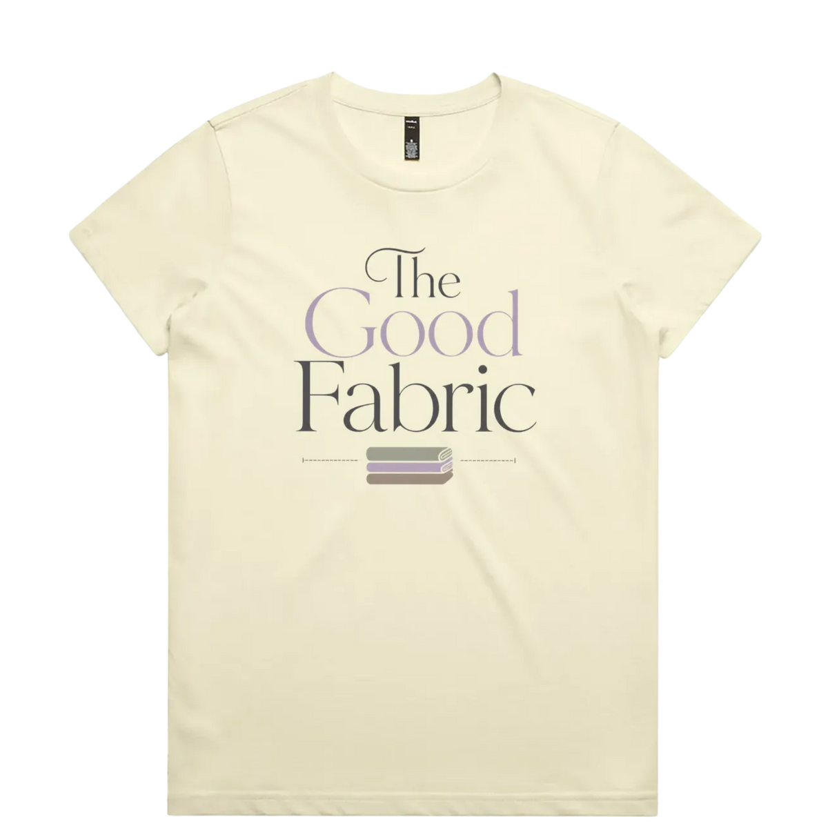 A very pale yellow short-sleeve crewneck t-shirt featuring a graphic print on the front that reads 'The Good Fabric' in dark gray and lavender lettering, with a small fabric bundle illustration below.