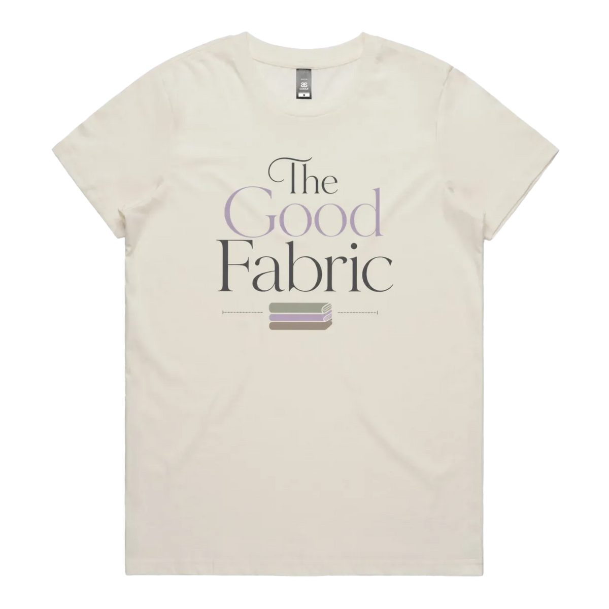 A pale cream short-sleeve crewneck t-shirt featuring a graphic print on the front that reads 'The Good Fabric' in dark gray and lavender lettering, with a small fabric bundle illustration below.
