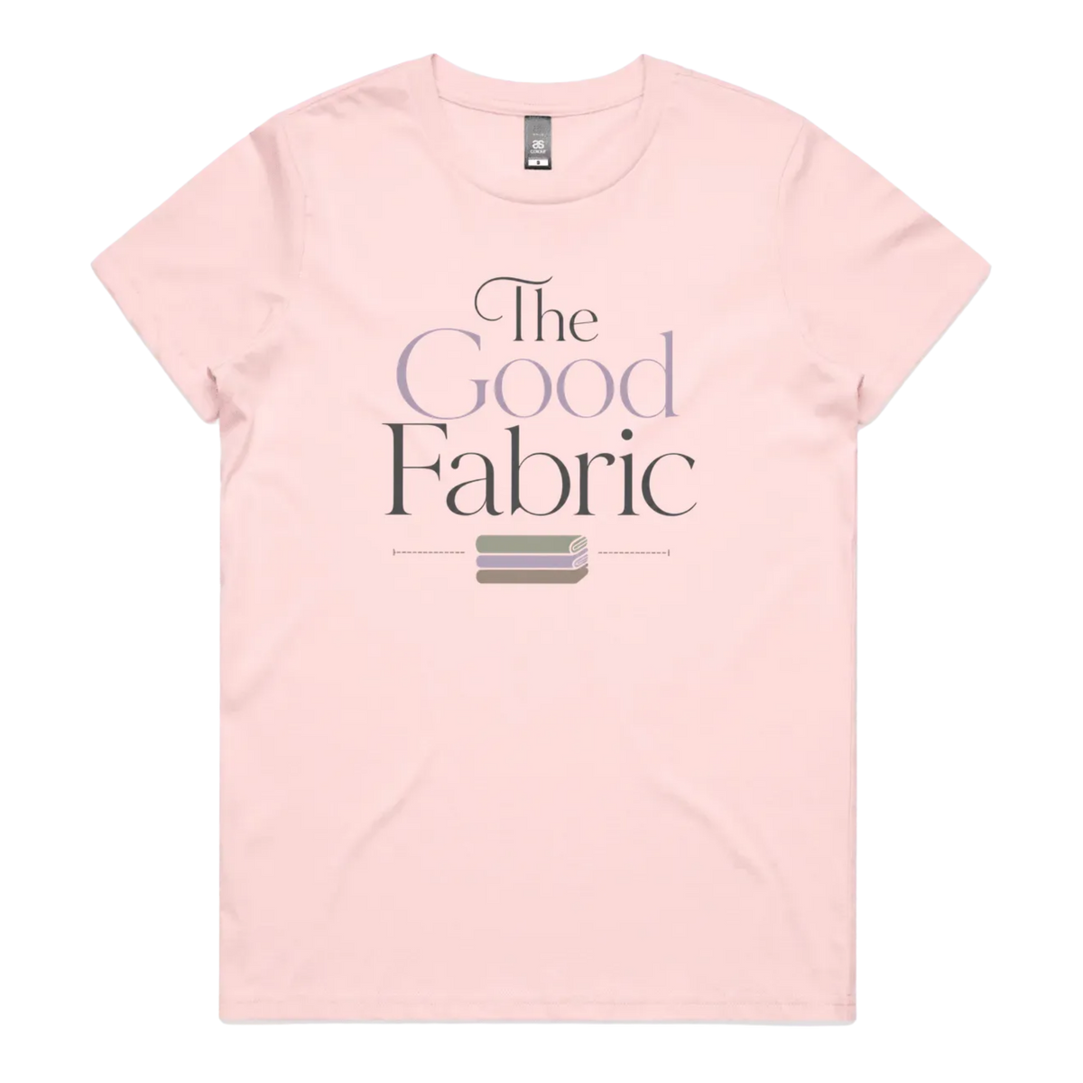 A pale pink short-sleeve crewneck t-shirt featuring a graphic print on the front that reads 'The Good Fabric' in dark gray and lavender lettering, with a small fabric bundle illustration below.