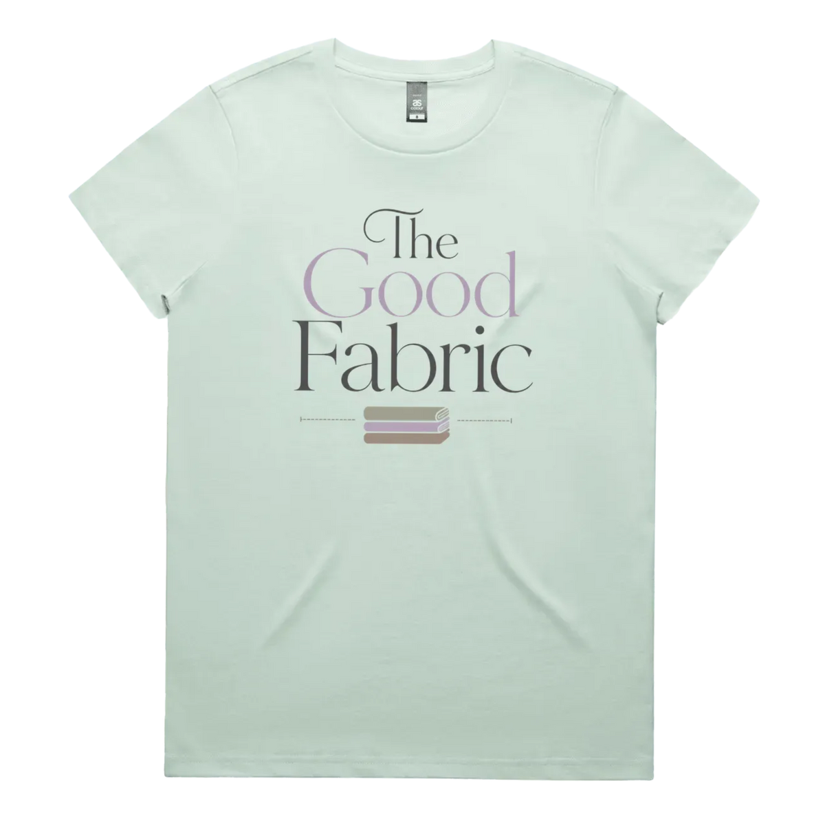 A pale green short-sleeve crewneck t-shirt featuring a graphic print on the front that reads 'The Good Fabric' in dark gray and lavender lettering, with a small fabric bundle illustration below.