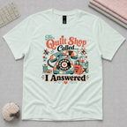 A light seafoam green graphic t-shirt laid flat on a pale grey cutting mat, surrounded by quilting props. The shirt features a colorful retro-style print of a patchwork covered rotary phone, shopping bags, and the text 'The Quilt Shop Called, I Answered'.