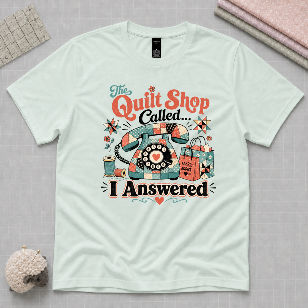 A light seafoam green graphic t-shirt laid flat on a pale grey cutting mat, surrounded by quilting props. The shirt features a colorful retro-style print of a patchwork covered rotary phone, shopping bags, and the text 'The Quilt Shop Called, I Answered'.