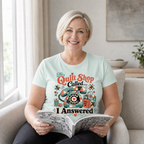 A smiling blonde woman sits in a light beige armchair, holding an open book. She is wearing a pale seafoam green graphic t-shirt with a retro-style patchwork covered telephone and the text 'The Quilt Shop Called, I Answered'.