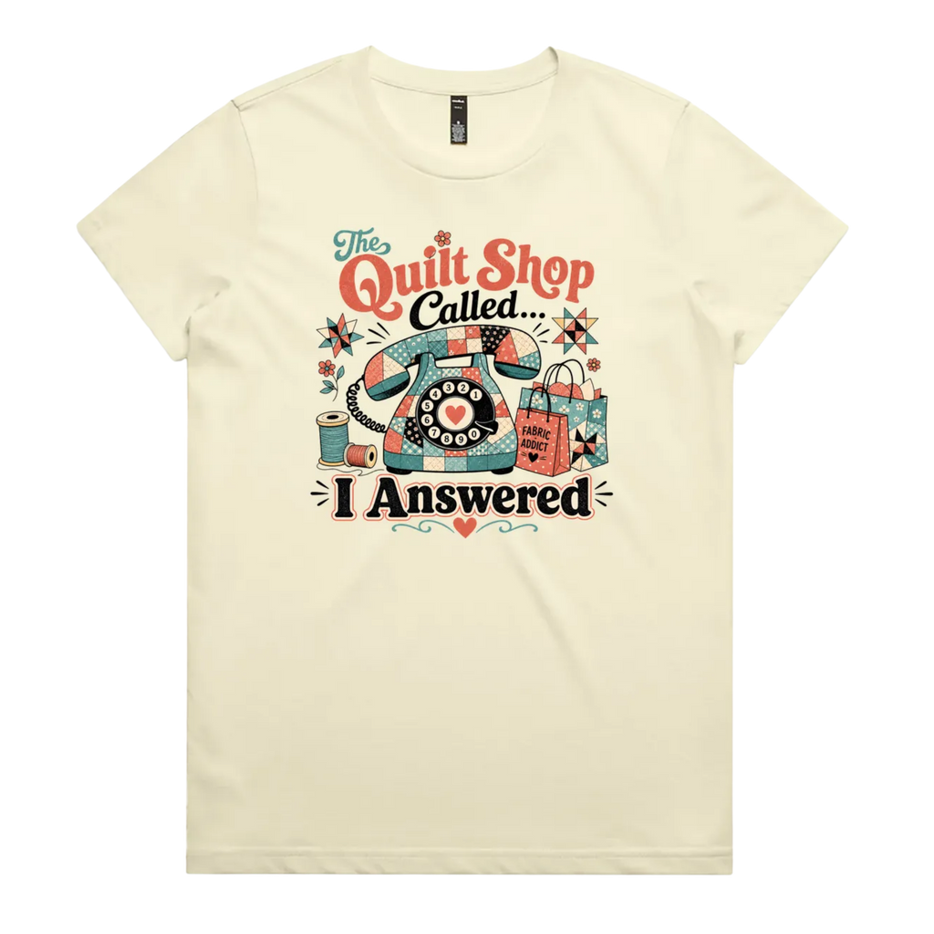 A pale butter yellow short-sleeve t-shirt with a colorful retro graphic on the chest featuring a vintage rotary phone made of fabric pieces, a spool of thread, shopping bags, and the text 'The Quilt Shop Called, I Answered'.