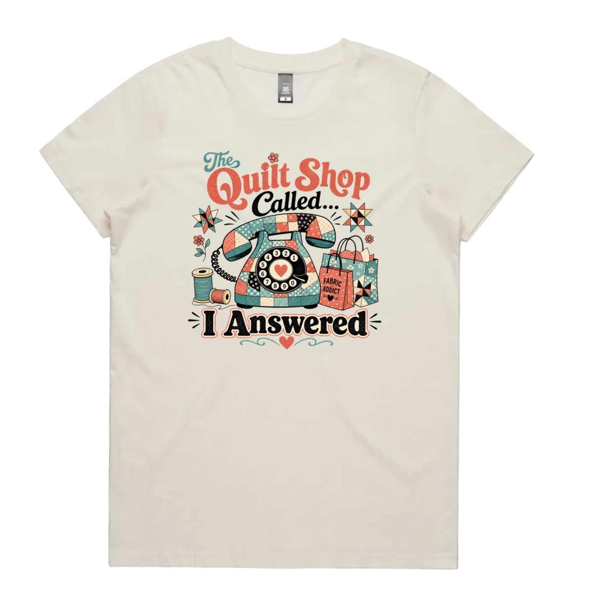 A light cream short-sleeve t-shirt with a colorful retro graphic on the chest featuring a vintage rotary phone made of fabric pieces, a spool of thread, shopping bags, and the text 'The Quilt Shop Called, I Answered'.
