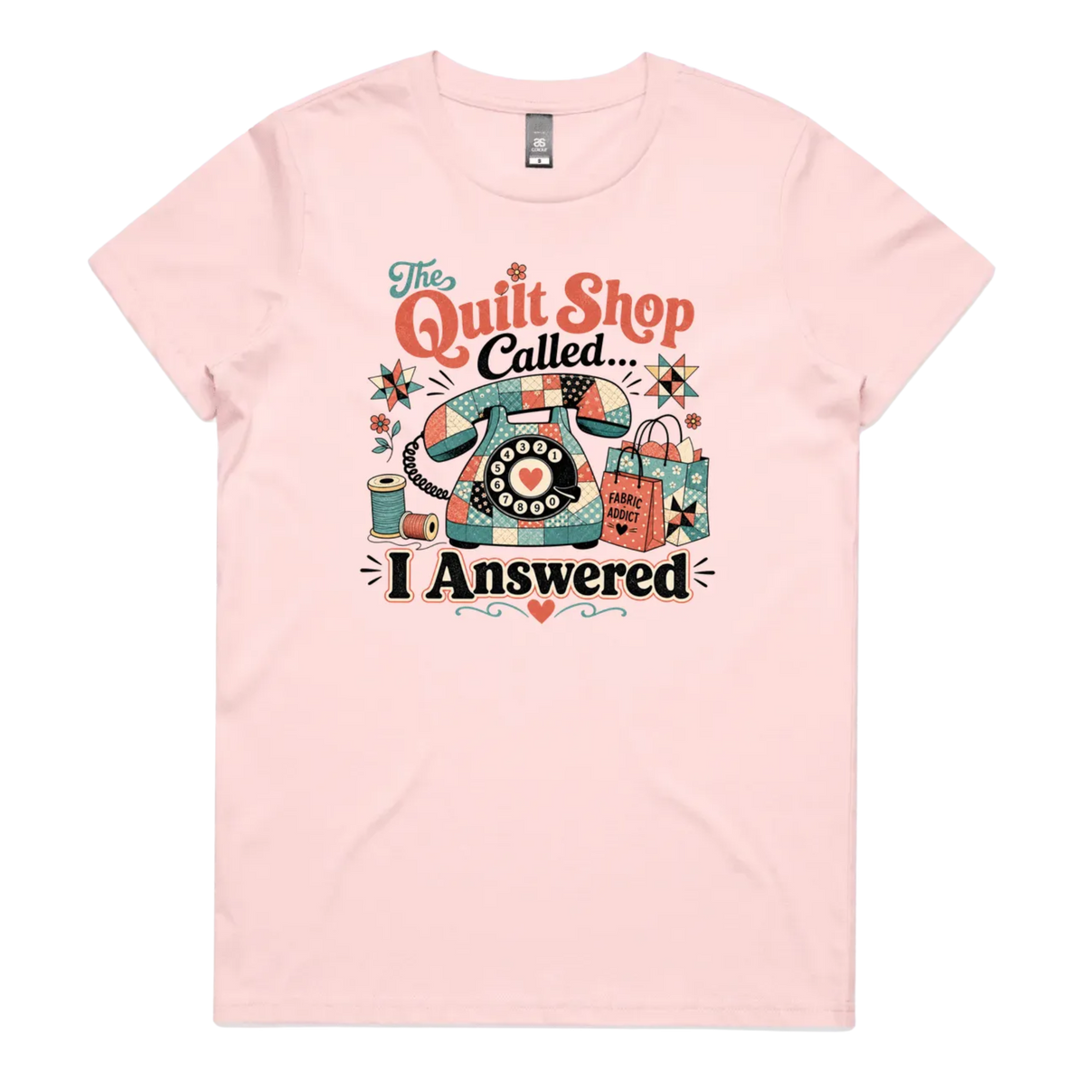 A light pink short-sleeve t-shirt with a colorful retro graphic on the chest featuring a vintage rotary phone made of fabric pieces, a spool of thread, shopping bags, and the text 'The Quilt Shop Called, I Answered'.