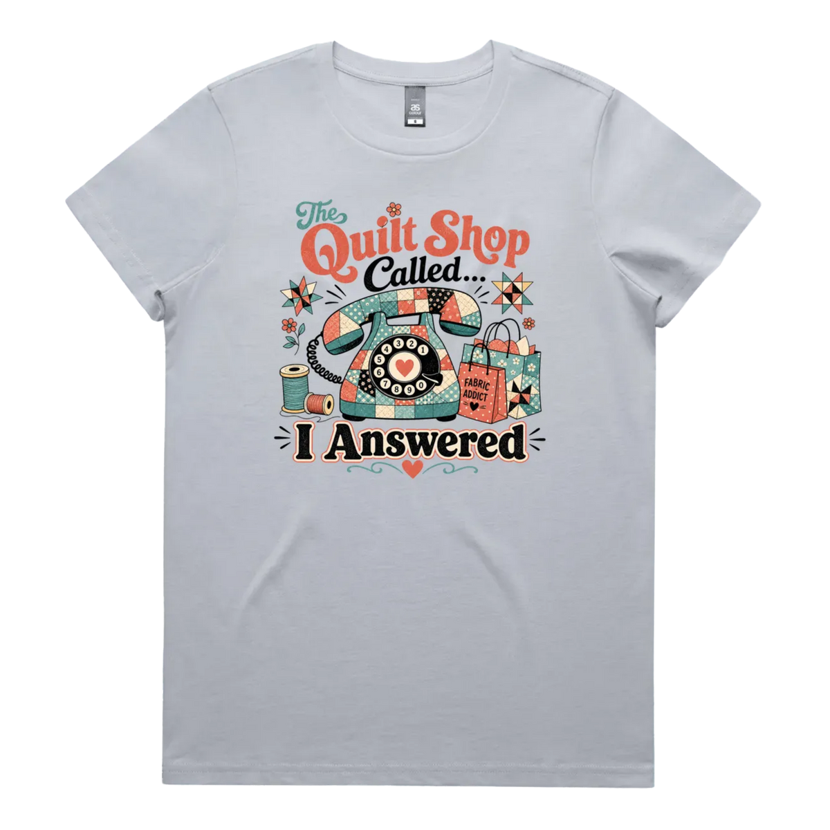 A light powder blue short-sleeve t-shirt with a colorful retro graphic on the chest featuring a vintage rotary phone made of fabric pieces, a spool of thread, shopping bags, and the text 'The Quilt Shop Called, I Answered'.