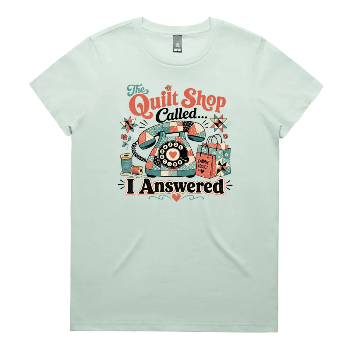 A light seafoam green short-sleeve t-shirt with a colorful retro graphic on the chest featuring a vintage rotary phone made of fabric pieces, a spool of thread, shopping bags, and the text 'The Quilt Shop Called, I Answered'.