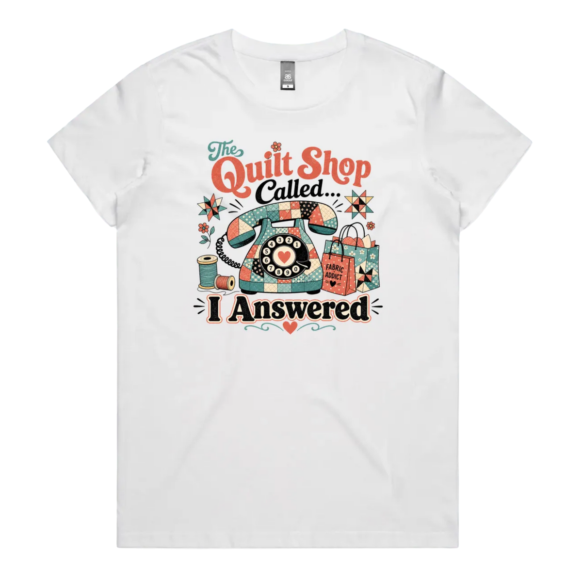 A white short-sleeve t-shirt with a colorful retro graphic on the chest featuring a vintage rotary phone made of fabric pieces, a spool of thread, shopping bags, and the text 'The Quilt Shop Called, I Answered'.