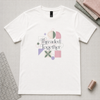 A pale cream short-sleeve crewneck t-shirt with a colorful graphic print reading 'Threaded Together' surrounded by sewing-themed shapes and threads, laid flat on a gray grid cutting mat with fabric and spools of thread around it.