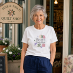 An older woman stands in front of a quilt shop doorway wearing a white graphic T-shirt with the phrase 'Threaded Together' and quilt-inspired geometric shapes, paired with navy drawstring pants. A 'Pine & Patch Quilt Shop' sign is visible behind her.