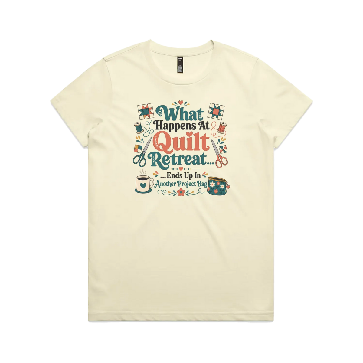 A pale butter yellow short-sleeve graphic t-shirt with the words 'What Happens at Quilt Retreat...ends up in another project bag'. The words are surrounded by quilt blocks thread spools, scissors, a coffee mug and zippered bag.