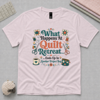 A pale mauve short-sleeve t-shirt laying flat on a pale grey cutting mat with quilting tools in the corners. The graphic on the t-shirt reads 'What Happends At Quilt Retreat...ends up in another project bag', surrounded by quilting tools.