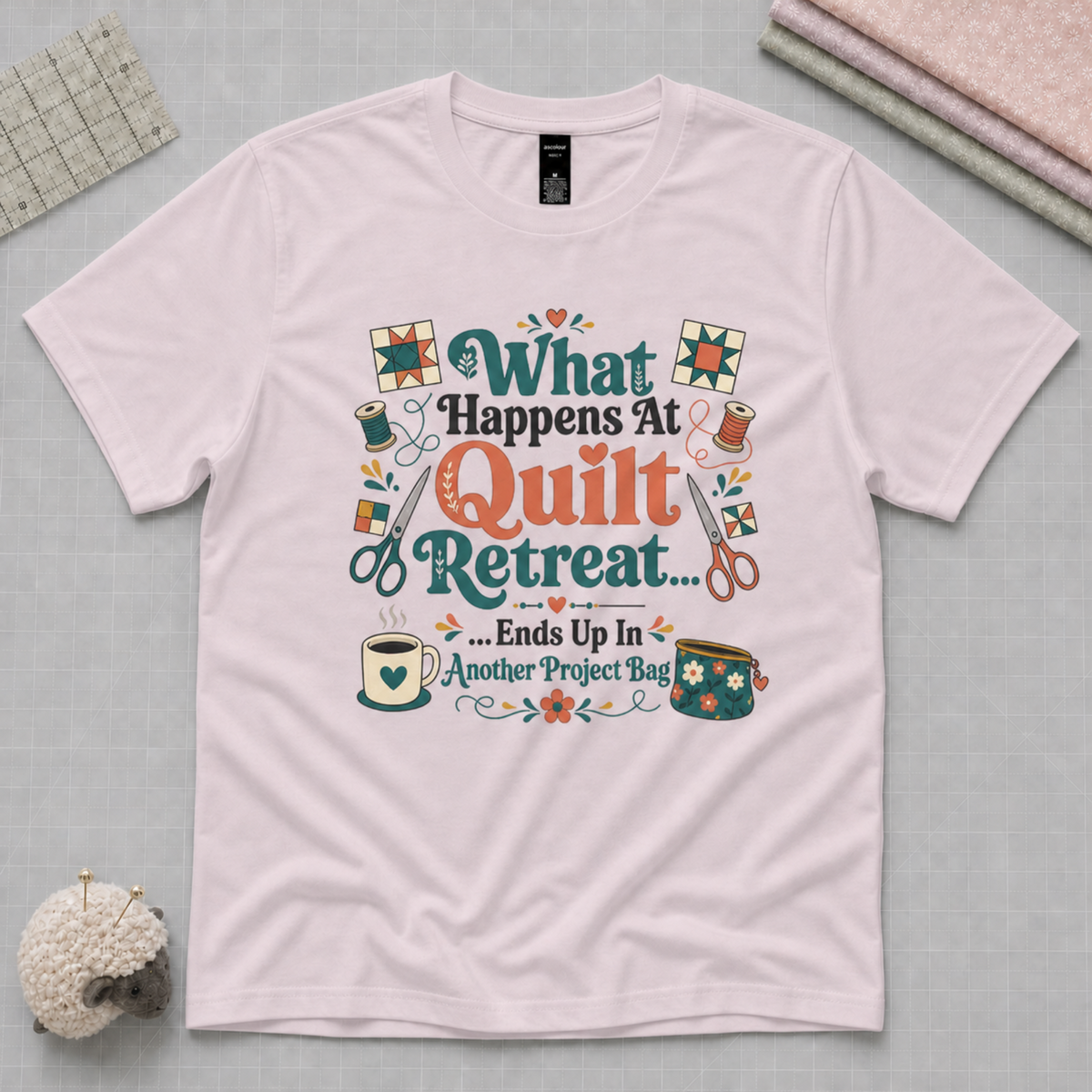 A pale mauve short-sleeve t-shirt laying flat on a pale grey cutting mat with quilting tools in the corners. The graphic on the t-shirt reads 'What Happends At Quilt Retreat...ends up in another project bag', surrounded by quilting tools.