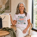 A woman with long grey hair sits at a table in a bright room wearing a pale mauve t-shirt with the graphic 'What Happends at Quilt Retreat...ends up in another project bag'. 