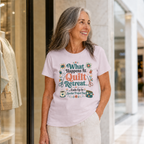A woman with long grey hair standing outside a boutique in a shopping mall wearing a pale mauve t-shirt. The graphic on the t-shirt reads 'What Happends At Quilt Retreat...ends up in another project bag', surrounded by quilting tools.