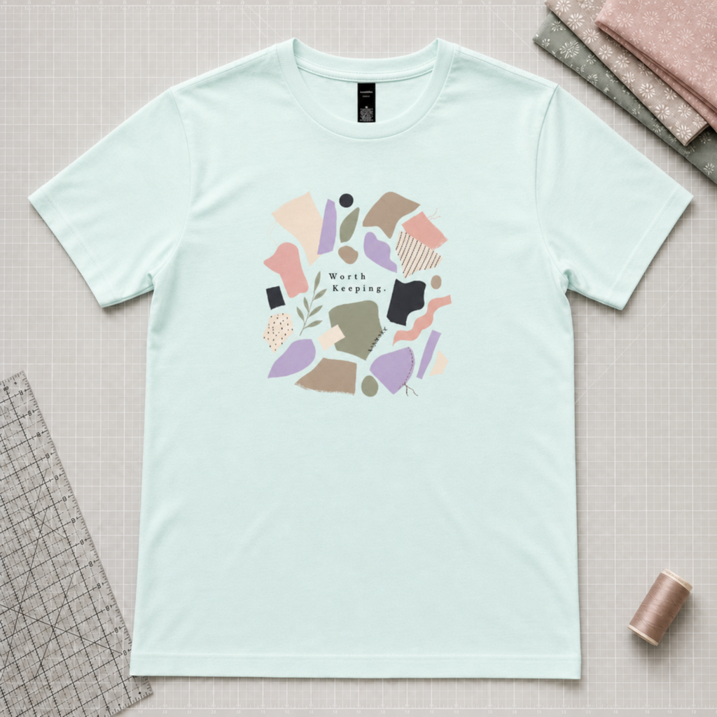 A light mint green short-sleeve t-shirt with a circular graphic print in the center featuring abstract shapes and the words 'World Keeping'. The shirt is laid flat on a gridded cutting mat surrounded by sewing and crafting props, including a ruler, spool of thread, and folded fabrics.