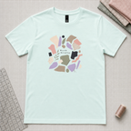 A light mint green short-sleeve t-shirt with a circular graphic print in the center featuring abstract shapes and the words 'World Keeping'. The shirt is laid flat on a gridded cutting mat surrounded by sewing and crafting props, including a ruler, spool of thread, and folded fabrics.