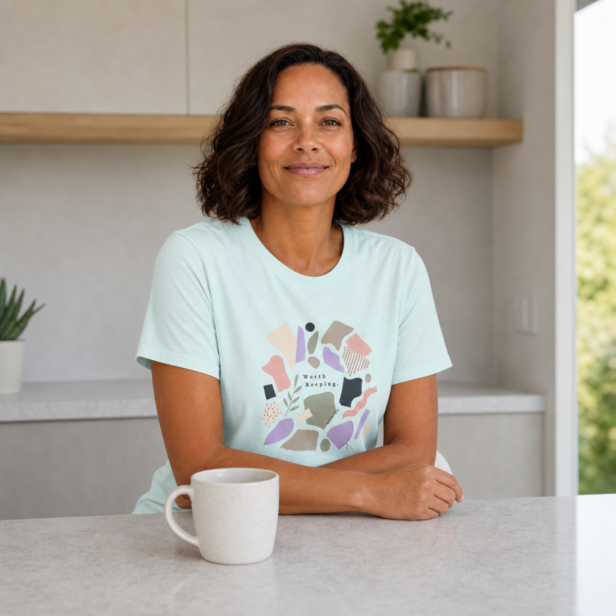 A woman with dark curly hair sits at a kitchen counter wearing a light mint green t-shirt with a colorful abstract graphic on the front with the words "Worth Keeping' surrounded by colourful fabric scraps in sage, mauve, cream. A white ceramic mug is in the foreground, with a modern kitchen interior and window in the background.