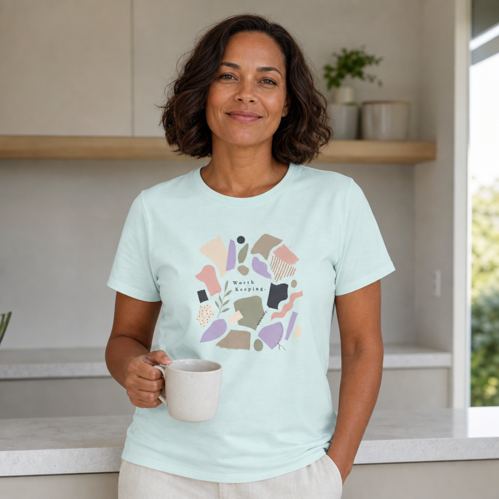 A woman wearing a light mint green graphic t-shirt with a colorful abstract design and the words 'Worth Keeping' holds a white ceramic mug in a bright kitchen setting.