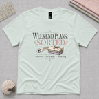 A light mint green graphic t-shirt laid flat on a gray cutting mat, with a quilting ruler, folded fabric and pincushion in the corners. The shirt features a graphic that reads 'WEEKEND PLANS: SORTED' above an illustration of a rotary cutter and tied bundle of fabric with 'fabric', 'friends', and 'sewing' underneath.
