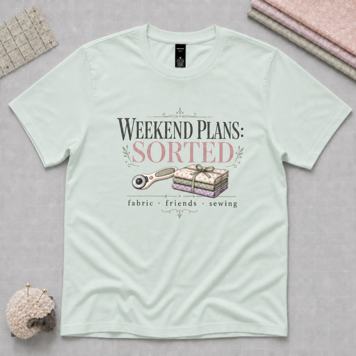 A light mint green graphic t-shirt laid flat on a gray cutting mat, with a quilting ruler, folded fabric and pincushion in the corners. The shirt features a graphic that reads 'WEEKEND PLANS: SORTED' above an illustration of a rotary cutter and tied bundle of fabric with 'fabric', 'friends', and 'sewing' underneath.