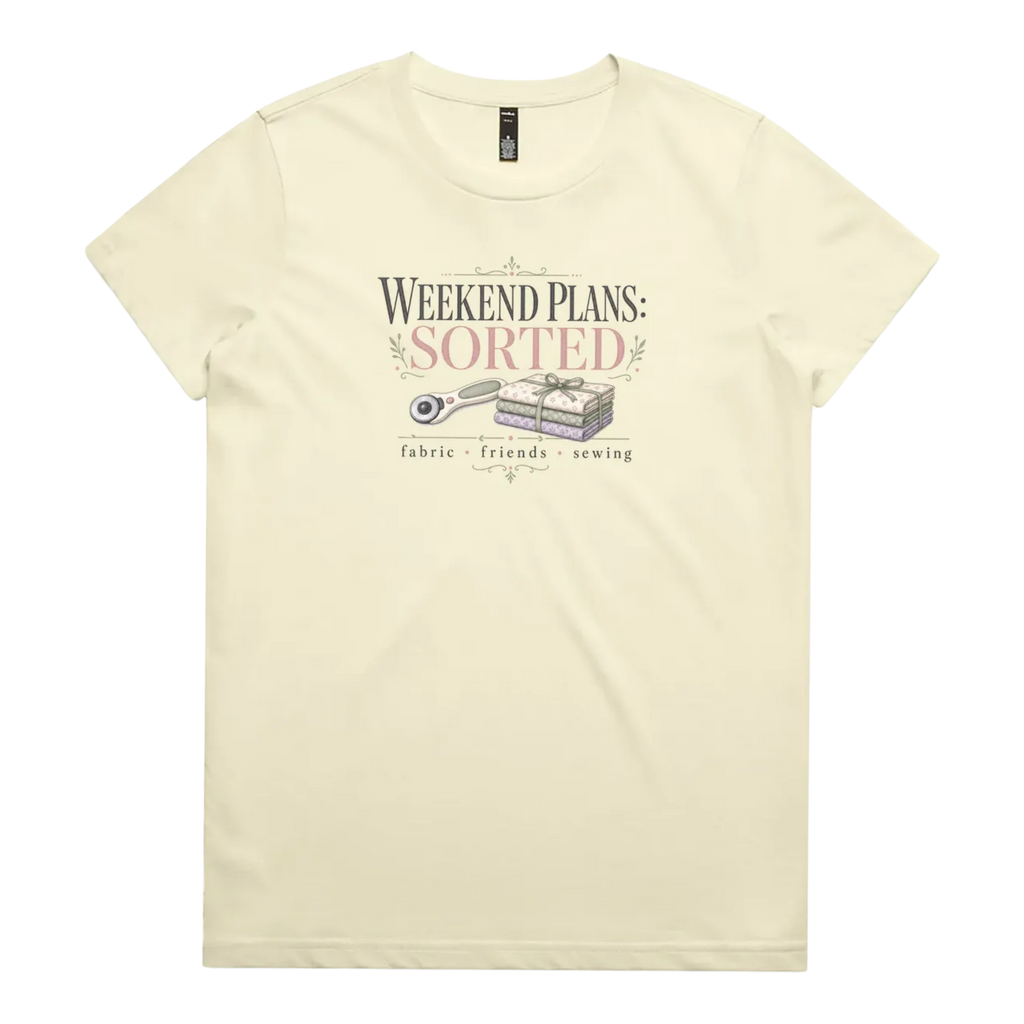 A pale butter yellow short-sleeve t-shirt with a vintage-style graphic on the chest reading 'WEEKEND PLANS: SORTED' above an illustration of a rotary cutter and tied fabric bundle, with 'fabric + friends + sewing' below.