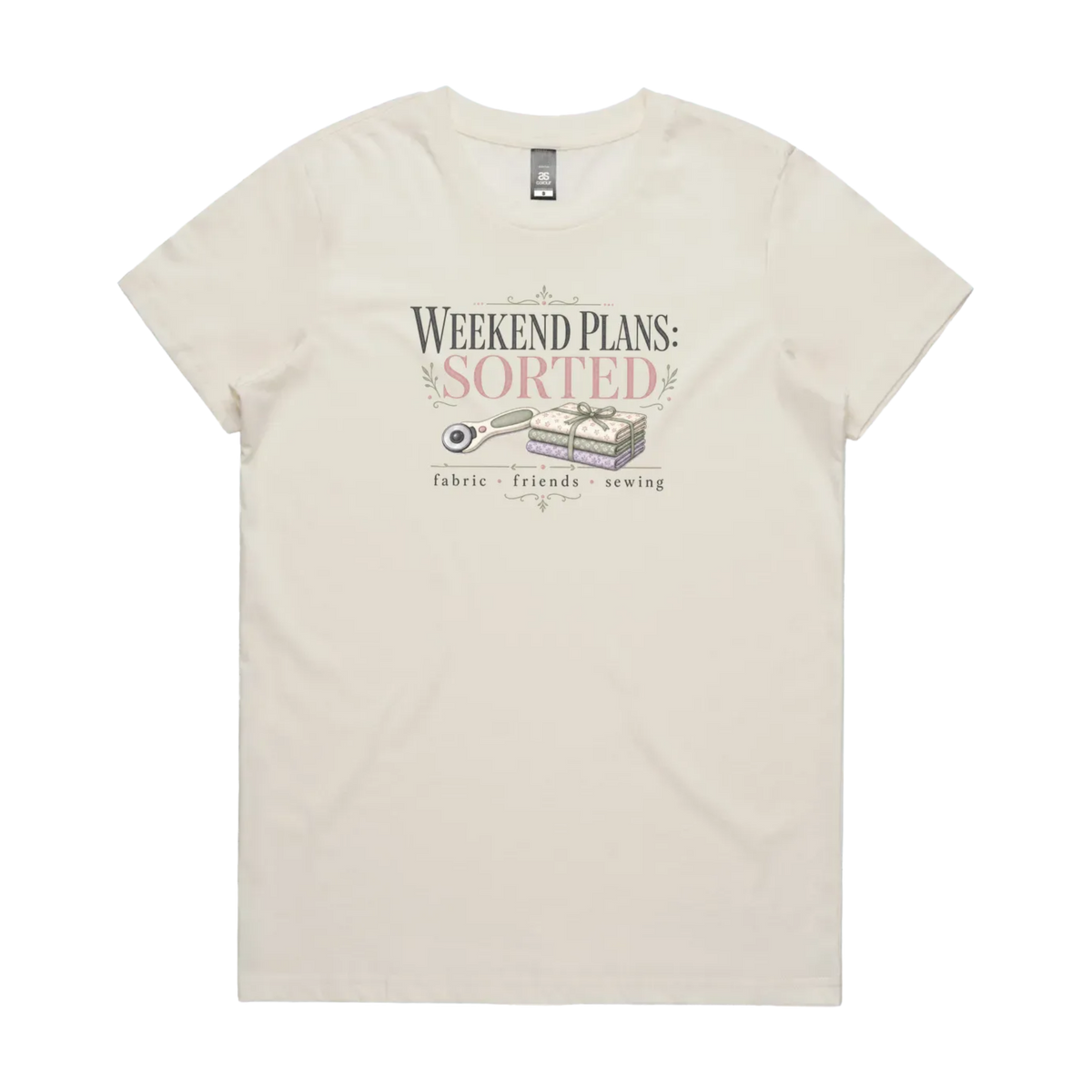 An off-white short-sleeve t-shirt with a vintage-style graphic on the chest reading 'WEEKEND PLANS: SORTED' above an illustration of a rotary cutter and tied fabric bundle, with 'fabric + friends + sewing' below.