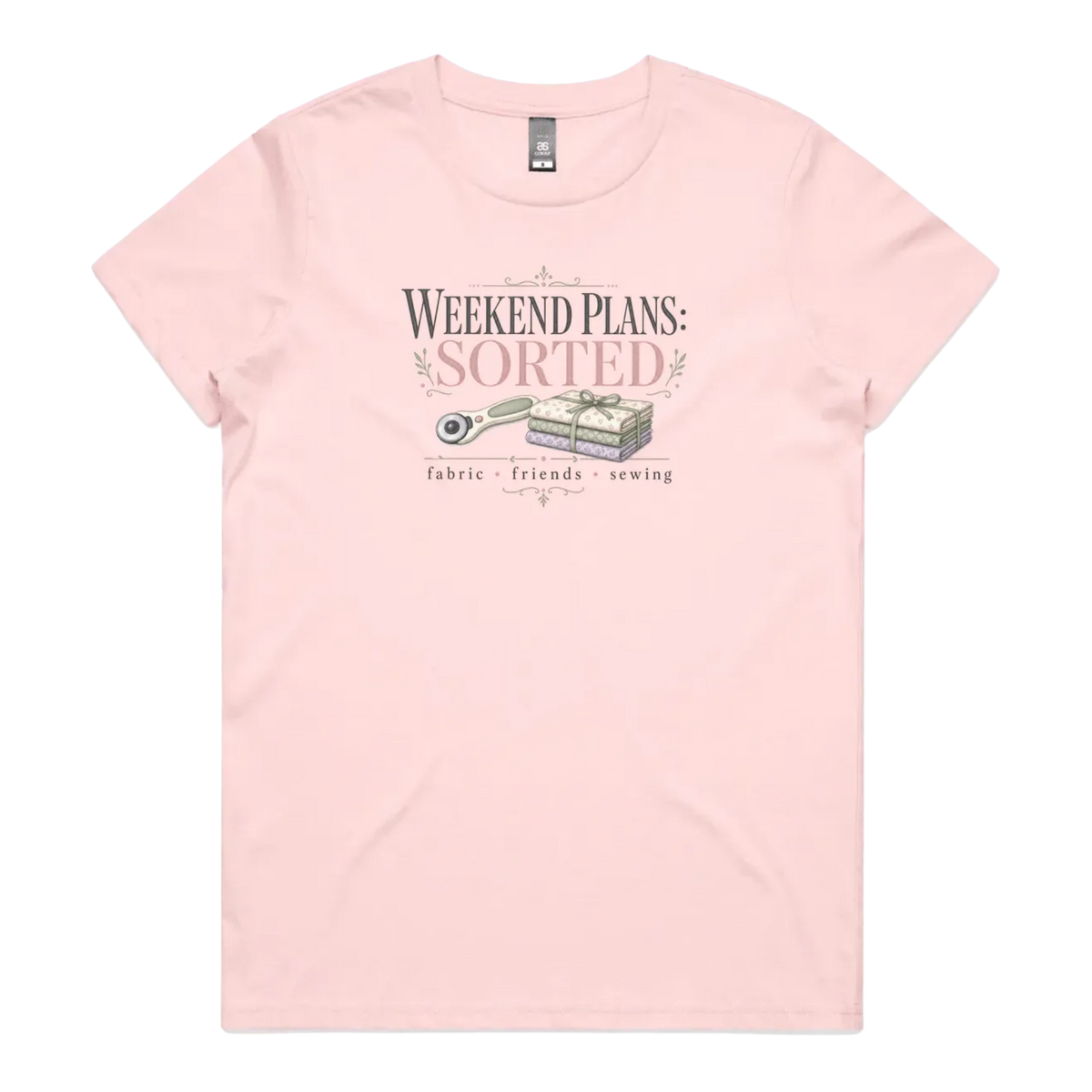 A light pink short-sleeve t-shirt with a vintage-style graphic on the chest reading 'WEEKEND PLANS: SORTED' above an illustration of a rotary cutter and tied fabric bundle, with 'fabric + friends + sewing' below.